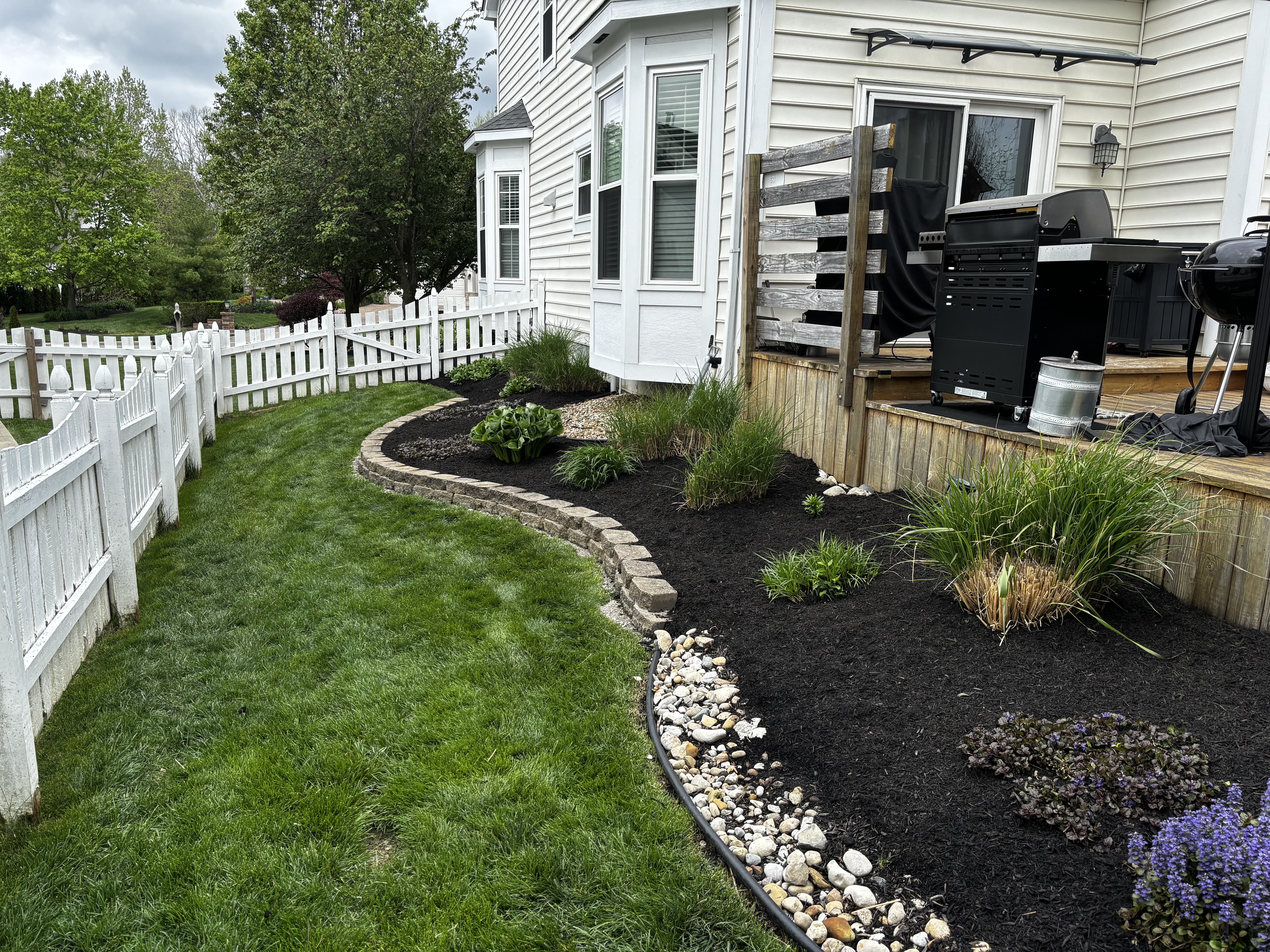 Curved stone retaining wall bordering a clean black mulched garden bed with perennial plants beside a wooden deck